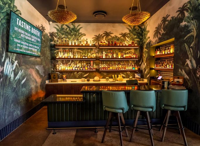 A Tasting Booth area with tropical leaf wallpaper, backlit spirit shelves, and teal bar stools.