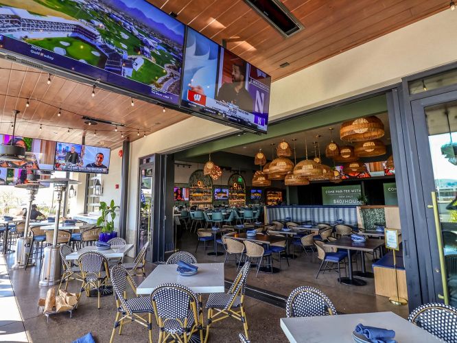 View from a patio looking into a vibrant restaurant/bar area with large TV screens and woven pendant lights.