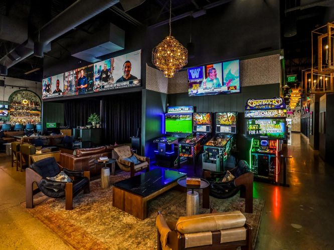 A lounge area with leather armchairs, a large rug, and arcade games like Golden Tee and Big Buck Hunter.