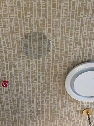 Close-up of patterned wallpaper on a ceiling with a circular vent and a portion of a gold light fixture.