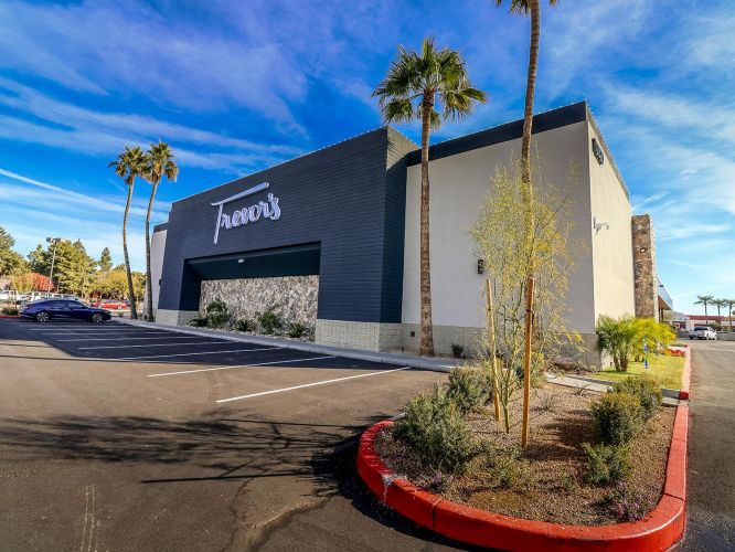 Wide exterior view of the Trevor's storefront with palm trees, landscaping, and clear blue skies.