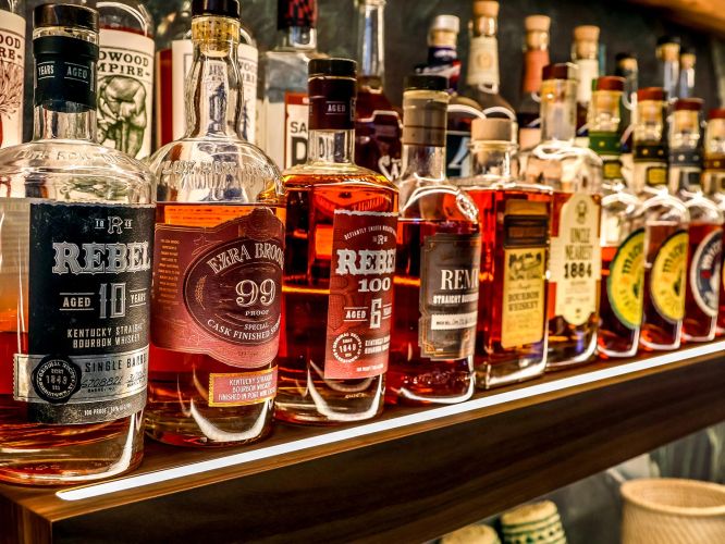 Close-up of various bourbon and whiskey bottles, including Rebel 10 and Ezra Brooks, on a glowing shelf.