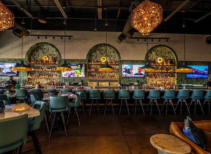 A long, stylish bar with teal stools, gold-accented shelving, and arched back-bar displays with tropical wallpaper.